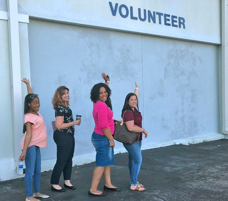 quatro mulheres apontando para uma placa de trabalho voluntário